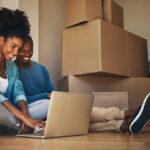 Couple staring at laptop on the floor with cardboard boxes next to them.